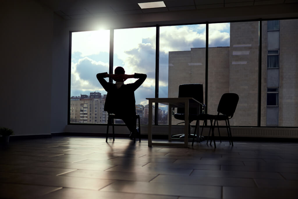 Գործնական կոստյումով ժամանակակից գրասենյակում գտնվող տղամարդը ձեռքերը բարձրացրել է գլխի հետևից՝ նայելով պատուհանից դեպի քաղաք: