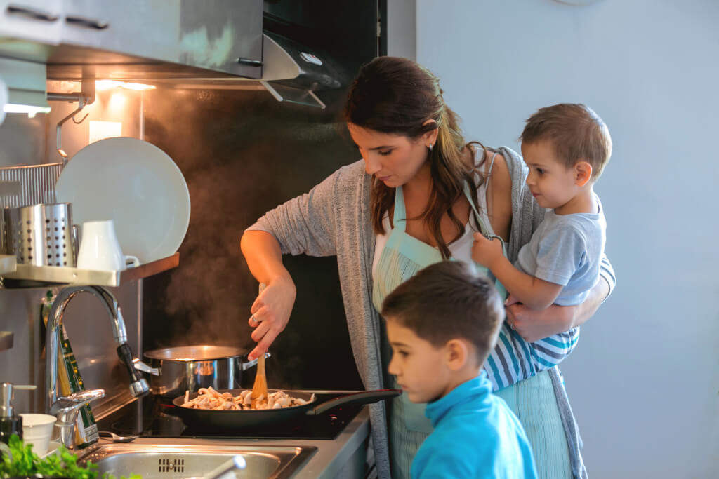 Mother holding toddler and cooking, older son standing