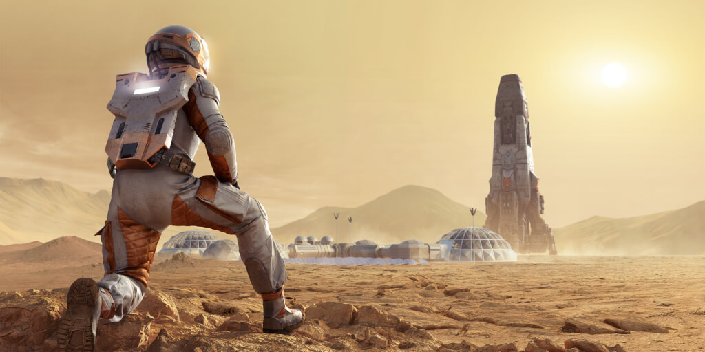 A close up image of an astronaut on Mars kneeling and looking at a rocket in the distance. The spaceman or spacewoman is dressed in full space suit viewed from behind, kneeling on rocks and looking into the distance at Mars base camp and rocket in the distance.