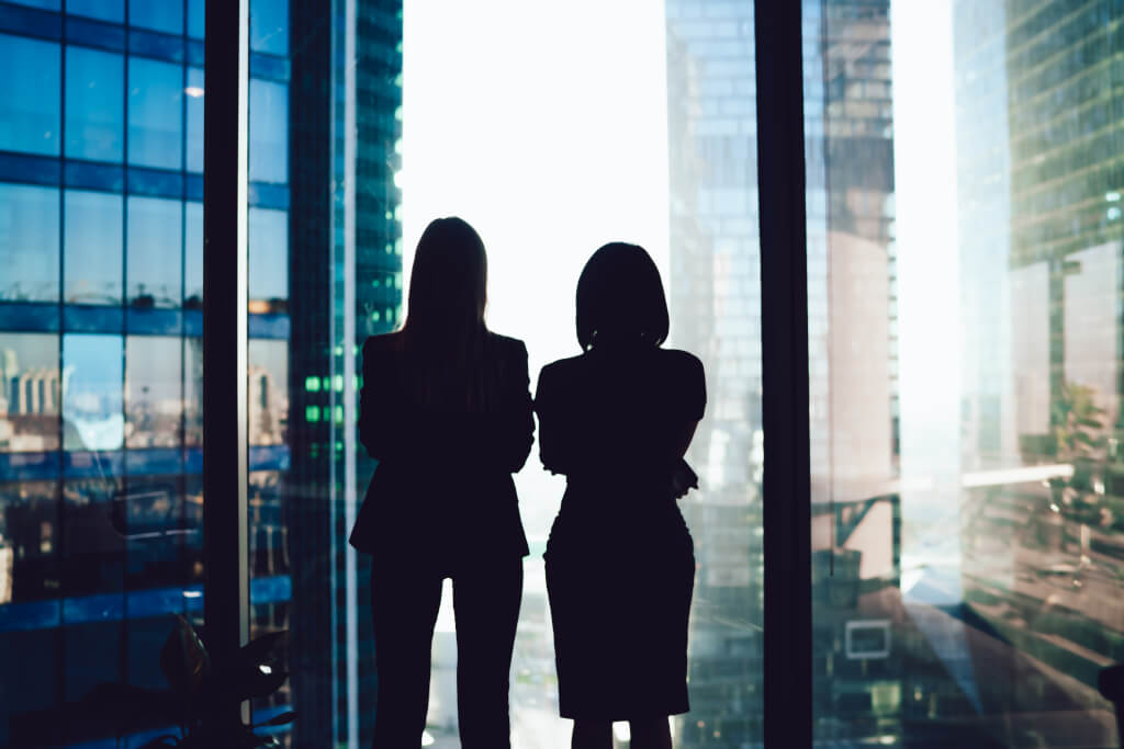 Back view of female colleagues in formal wear standing near window looking at modern exterior of skyscrapers in business center, silhouette of women