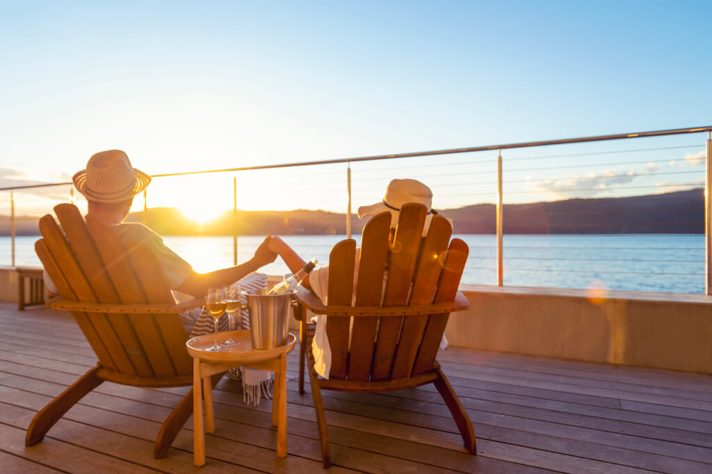 Couple relaxing and drinking wine and holding hands on deck chairs in an over water bungalow. They are looking at the view