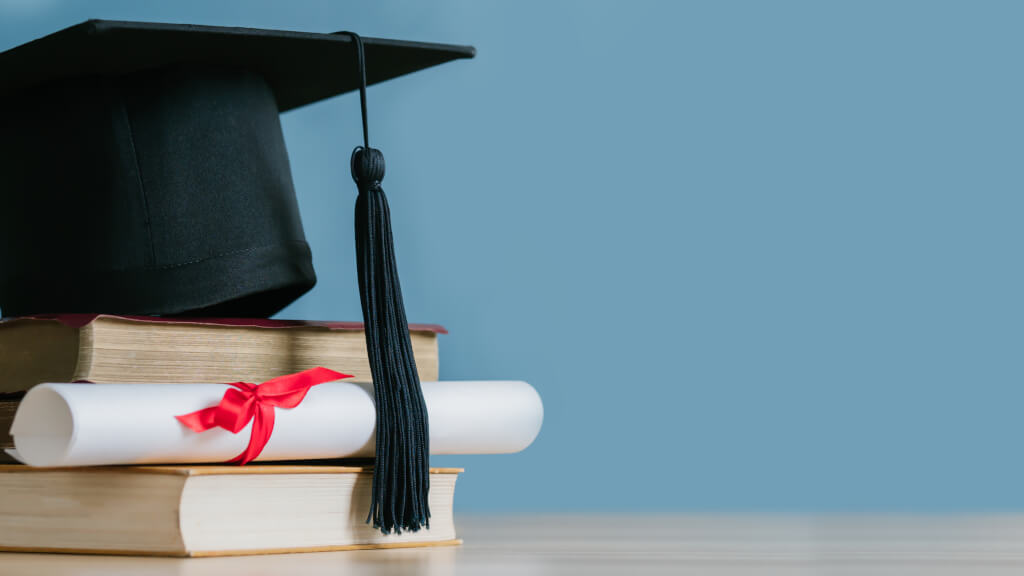 A mortarboard and graduation scroll on stack of books with blue background