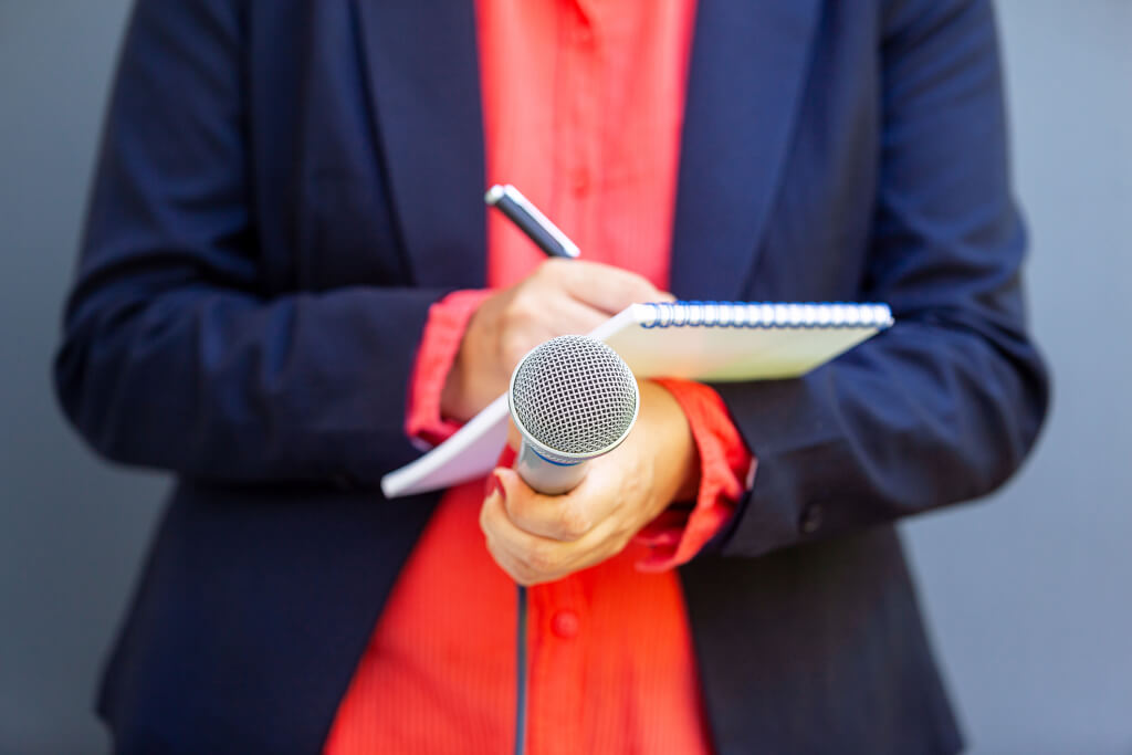 News reporter or TV journalist at press conference or media event, holding microphone and writing notes. Broadcast journalism concept.